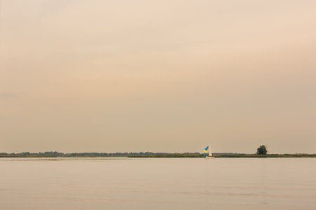 A peaceful scene of a sailboat gliding on a tranquil lake with a soft, warm-toned sky and distant greenery.の写真素材