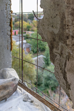 A damaged concrete wall with exposed rusty reinforcement wires, overlooking a blurry residential area with trees and rooftops in the background.の写真素材
