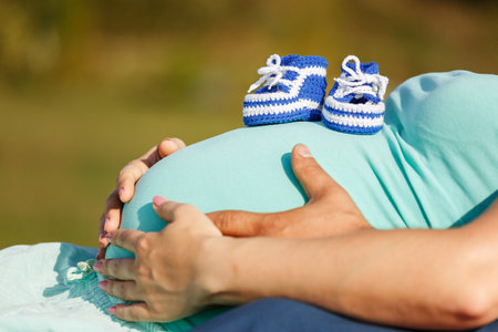 Pregnant mother on the spring meadow, lying on the green grass caresses her belly, close-up. Shoes for a newborn baby on the mother's stomach. Concept of healthy pregnancy, expectant mother outdoors.の写真素材
