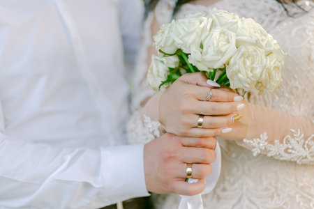 Close-up of bride and groom holding a bouquet of white roses, showing their wedding rings and elegant attire on their special day.の写真素材