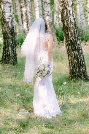 The bride in a white lace dress with a long veil holds a bouquet of flowers, outdoors, standing with her back. Summer weddingの写真素材