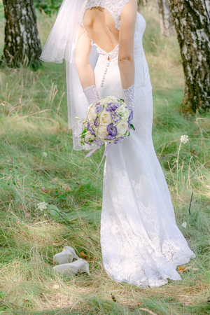 The bride in a white lace dress with a long veil holds a bouquet of flowers, outdoors, standing with her back. Summer weddingの写真素材