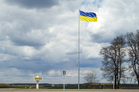 A large Ukrainian flag flutters on a tall flagpole against a cloudy sky, with a rural landscape and road sign in the background.の写真素材