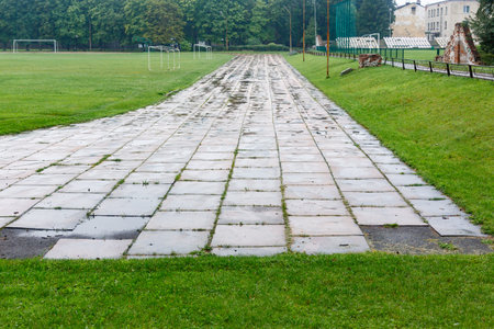 Old damaged running track in stadium with faded white lines and broken tiles, surrounded by green grass and fields. Urban sports facilities concept.の写真素材