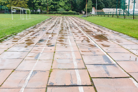 Old damaged running track in stadium with faded white lines and broken tiles, surrounded by green grass and fields. Urban sports facilities concept.の写真素材