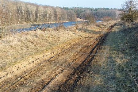 A rugged, muddy dirt road runs alongside a calm river, surrounded by dry grass and bare trees in a peaceful countryside setting.の写真素材