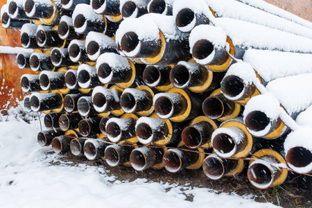 Stack of industrial insulated pipes covered with fresh snow in a residential construction area during heavy winter snowfall.の写真素材