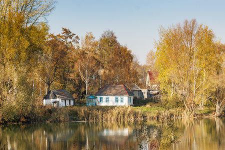 Traditional village houses surrounded by autumn trees reflect in a peaceful river, creating a serene and picturesque countryside landscape.の写真素材