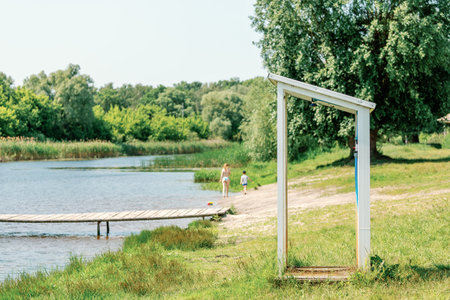 Public outdoor shower on grassy lake beach with people relaxing and playing near water on a sunny summer day.の写真素材