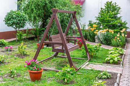 A wooden swing bench stands in a landscaped backyard garden with potted flowers, green shrubs, and blooming orange lilies.の写真素材