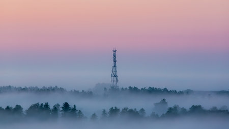 A communication tower rises through dense morning fog above a forest, with pastel-colored sky...