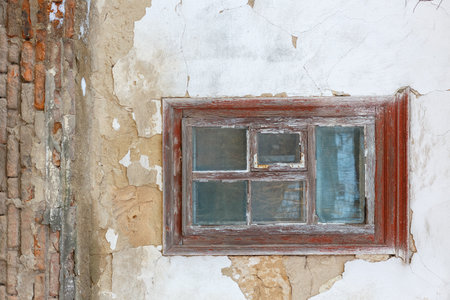 Rustic wooden-framed window with peeling paint set in a cracked, weathered plaster and brick wall of...