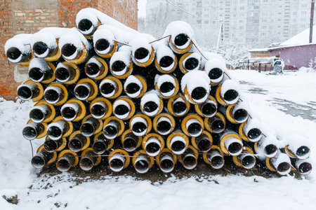 Stack of industrial insulated pipes covered with fresh snow in a residential construction area during heavy winter snowfall.の写真素材