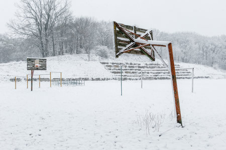 Old basketball hoops stand in an empty outdoor court, surrounded by snow-covered trees and bleachers during a cold winter day.の写真素材