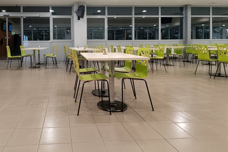 A spacious, brightly lit cafeteria featuring rows of green chairs and white tables, creating a clean...