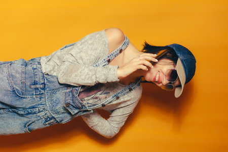 A cheerful woman in sunglasses, a denim cap and denim overalls smiles while talking on the phone. She poses against a bright background, giving the picture a casual and optimistic lookの写真素材