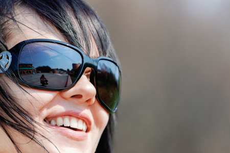 Close-up portrait of smiling woman with black hair wearing large sunglasses, with city reflection visible in the lenses on a sunny day.の写真素材