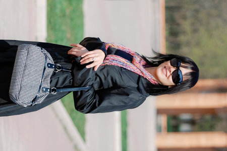 Portrait of fashionable woman wearing black coat, sunglasses and colorful scarf, holding handbag while walking in sunny city park.の写真素材