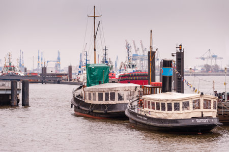 Hamburg, Germany - March 01, 2014: Two vintage barges lay at anchor at Museumshaven Oevelgoenne at hazy day. In the background of modern red harbourtugs, container terminal and cranes of the shipyard Blohm + Voss.のeditorial素材