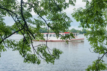 Looking through a courtain of branches of trees at Alster Lake in summer and Alster Tourist Ship in Hamburg, Germany in blurry background with selective focus.の写真素材
