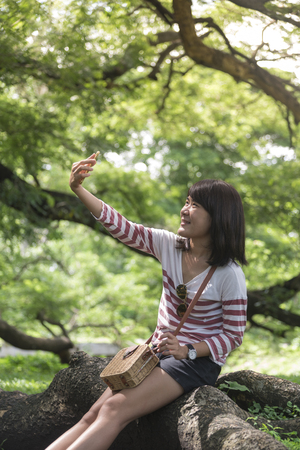 Beautiful young Asian woman taking a selfie with smart phone outdoors in summer in park. Horizontal image.の写真素材