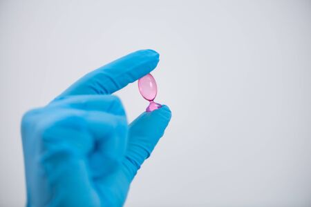 Young attractive female scientist with protective eyeglasses and mask holding a transparent pill with fingers in gloves in the pharmaceutical research laboratoryの写真素材