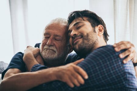 Sweet bonding. Cheerful elderly man sitting on the sofa next to his adult son, hugging him and...