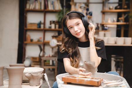 Beautiful young woman holding pottery instrument for scraping, smoothing, shaping and sculpting. Lady siting on bench with pottery wheel and making clay potの写真素材