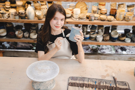 Beautiful young woman holding pottery instrument for scraping, smoothing, shaping and sculpting. Lady siting on bench with pottery wheel and making clay potの写真素材