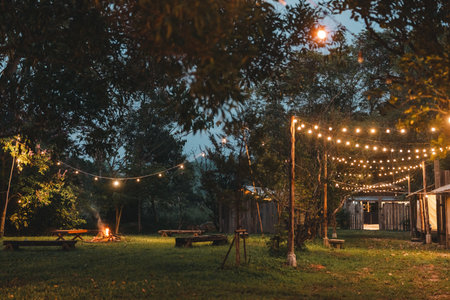 Cozy outdoor campfire surrounded by rustic wooden benches and glowing string lights, creating a peaceful evening atmosphere perfect for glamping under the stars in a nature-inspired forest retreatの写真素材
