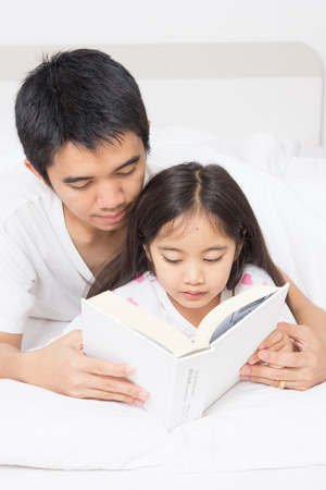 Little Asian child reading a story book with father on the bedの写真素材