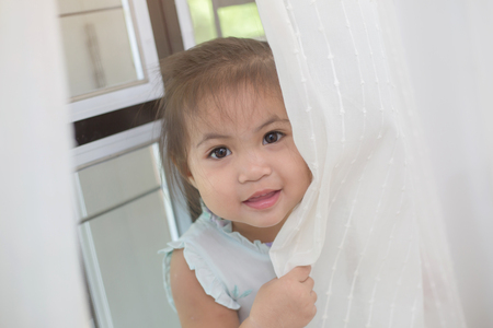 Asian girl playing hide and seek behind the curtainの写真素材
