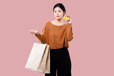 Happy Asian woman holding shopping bag and showing credit card isolated over pink background.の写真素材
