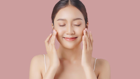 Young beautiful Asian woman massages her face gently with fingertips isolated over pink background. Face cream commercial advertising concept.の写真素材