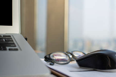 A laptop, mouse, eyeglasses, and notebook are placed on a table by a large window in the home office.の写真素材