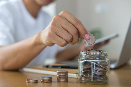 Saving money concept. Female hand putting money coin stack in the jar for the future, growing business, education and retirement on office desk at home. She felt that saving money made his life safe.の写真素材