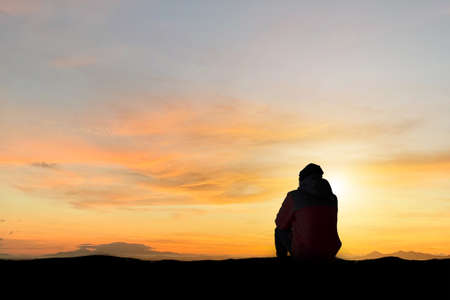 Silhouette of young man sitting and watching beautiful view overcast sky, sunset alone on top of the mountain. He enjoyed traveling and was successful when he reached the summit.の写真素材