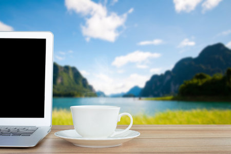 Coffee morning and laptop on wooden table on front of blurred background of the lake, mountain, meadow and blue sky among bright sunlight on a clear day.の写真素材