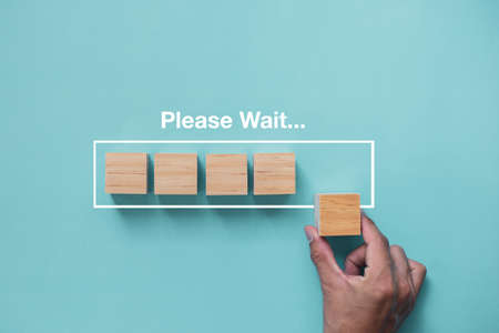 Close up image of hand putting wood cube in progress bar and please wait a moment written on the blue background.の写真素材