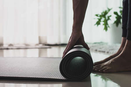 Young woman practicing yoga and meditating. Woman hand is rolling up and preparing exercise mat for practice online yoga class in the morning at home.の写真素材