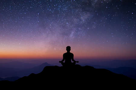 Silhouette of young female sitting practices yoga and meditating in lotus position alone on top of the mountain with night sky, star, Milky Way and meteor. She felt calm and happy.の写真素材