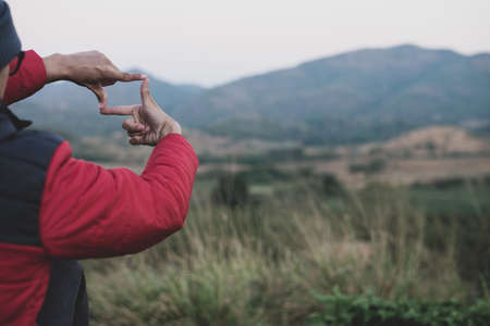 New year planning and vision concept, Young traveler wearing red sweater sitting alone on top of mountain hands making frame gesture and capturing beautiful view of meadow and mountain with copy spaceの写真素材