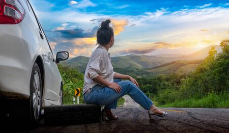 Young beautiful lady sit near car for calling for help on the public road in forest area at mountain and sky backgroundの写真素材