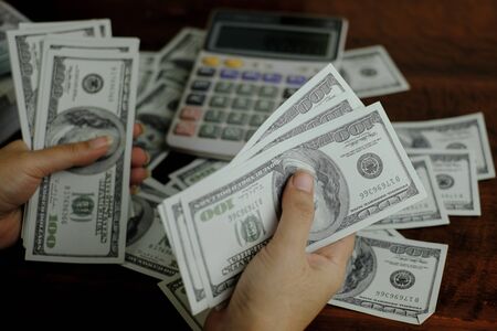 Hand holding smartphone and calculator on a stack of 100 US dollars banknotes lots of moneyの写真素材