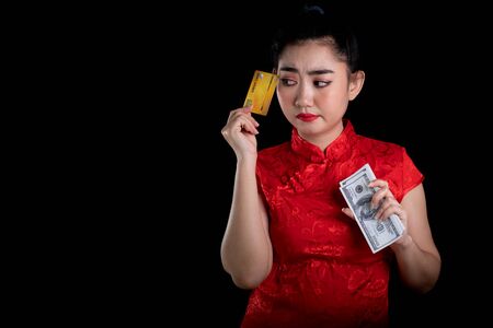 Portrait of a beautiful young Asia woman red dress traditional cheongsam holding credit card and money banknote 100 USD at the black background, China girl Chinese new year conceptの写真素材