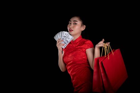 Portrait of a beautiful young Asia woman red dress traditional cheongsam holding bag shopping and money banknote 100 USD at the black background, China girl Chinese new year conceptの写真素材