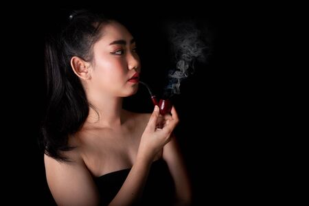 Portrait beautiful Asia young women with a smoking pipe tobacco on the black background, Studio shot close up Image of cigarette smoke spread in lady's handの写真素材