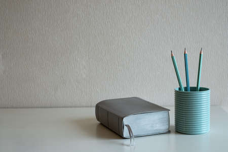 A book and pastel pencils in the blue glass on the desk at gray wall background