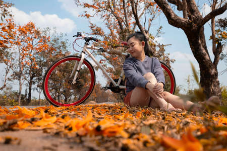 Beautiful young asia woman sitting next to her bike outdoors at palash tree with full of beautiful orange flower background, Butea Monosperma or Butea frondosa of southeast asia from natureの写真素材