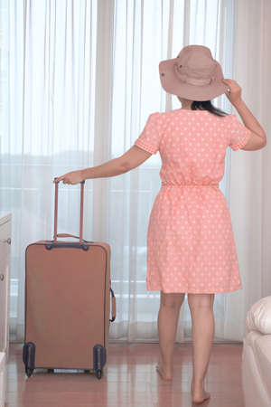 Asian tourist woman in a pink dress standing with her luggage in the hotel bedroom, Happy women lifestyle with holiday summer travel vacation conceptの写真素材